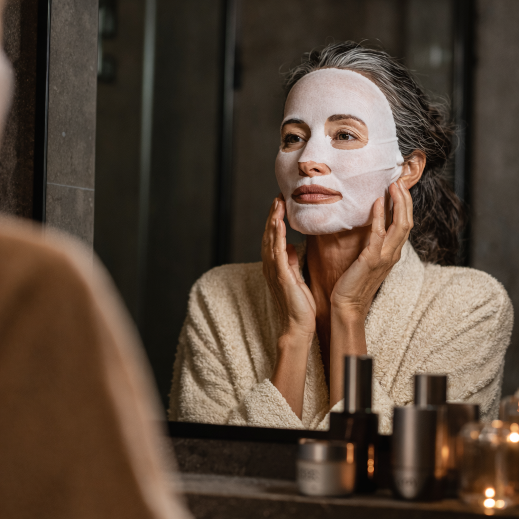 Woman applying a Merikit Medi Cell Coconut Gel Mask in front of a mirror with skincare products on a table.